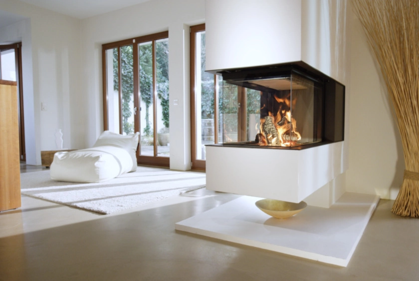 Modern vardagsrum med panoramafönster, en vit hörnspis med brasa och en ljus matta. Eldstadens eleganta design skapar en varm och inbjudande atmosfär.