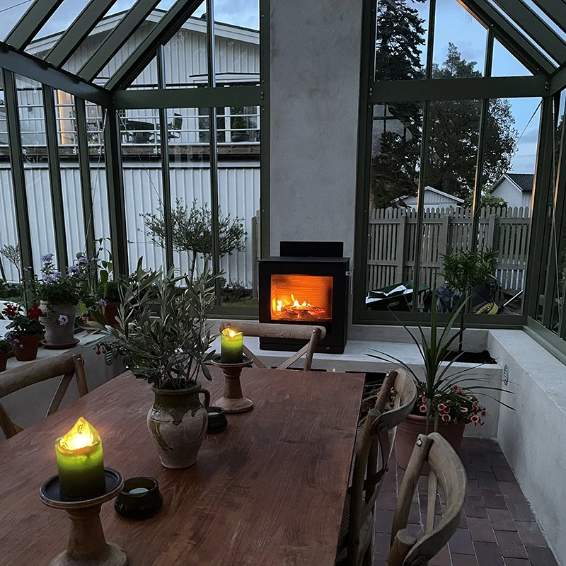 Inglasat uterum med träbord, tända ljus och en braskamin, omgiven av växter och stora fönster med utsikt mot en trädgård.