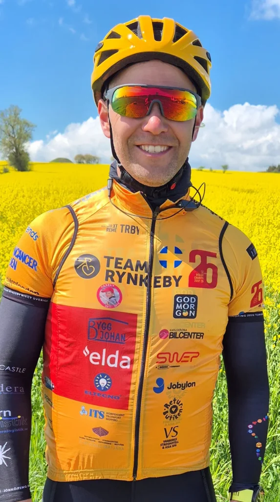 Cyklist i gul hjälm och Team Rynkeby-tröja står framför blommande rapsfält under klarblå himmel.