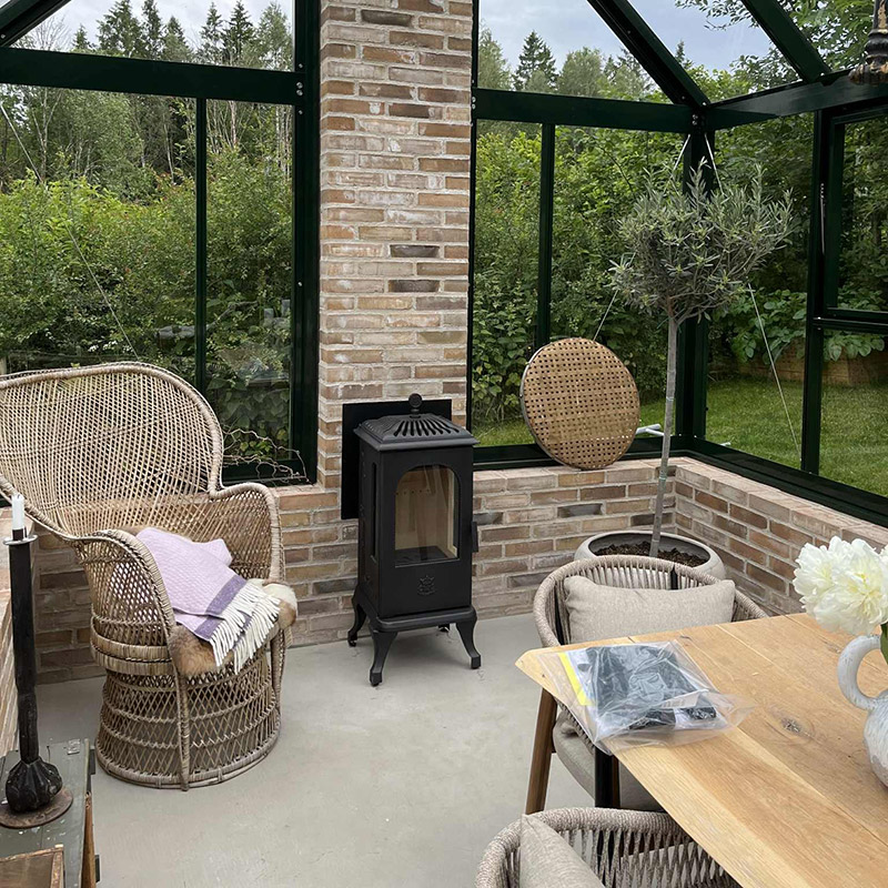 Inglasad uteplats med rottingmöbler, kamin och träbord. Växter omger rummet med tegelväggar, som ger en rustik känsla. Grönska syns genom glasfönstren.