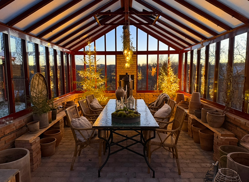 Inglasad veranda med juldekorationer, inklusive upplysta granar och belysning. Rustika möbler och blomkrukor omger ett långt bord. Solnedgång syns genom fönstren.
