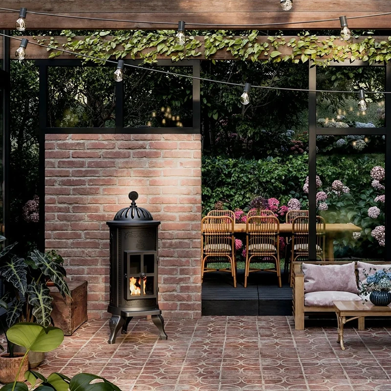 Uteplats med tegelvägg, kamin och belysning. Växter och blommor omger en matplats med rottingstolar. Trädgårdsinredning och utomhusmys.