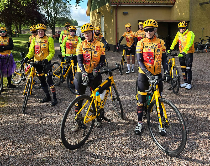 Cykelgrupp i gula tröjor och hjälmar står med sina cyklar på en grusplan framför en gul byggnad, redo för en cykeltur.