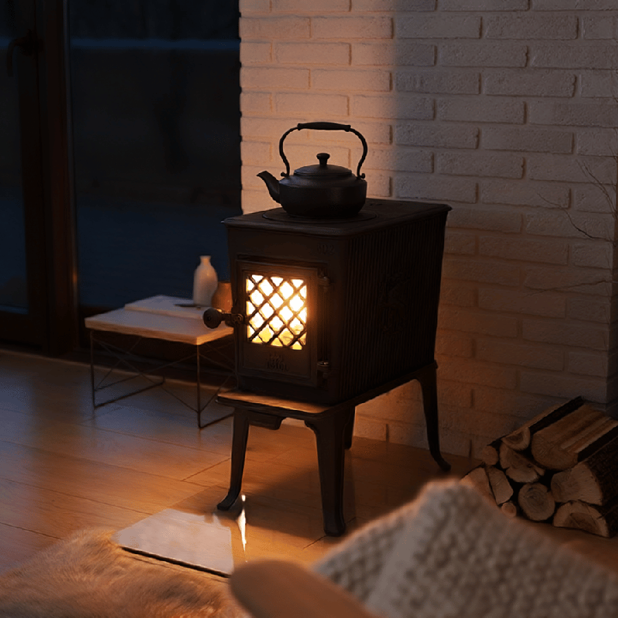 Vedeldad kamin med te-kanna på toppen, brinnande eld inuti, omgiven av vedträn och tegelvägg. Mysig atmosfär i ett vardagsrum.