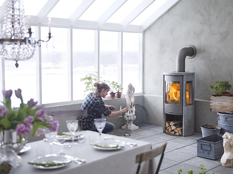 En person justerar en skulptur i ett ljust rum med stora fönster, en tänd kamin och en dukad matplats med blommor.