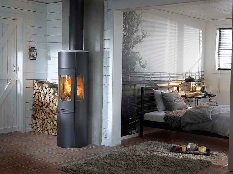 Modern kamin i hörn av ljus inredning med staplat vedträ. Intill ligger ett sovrum med dubbelsäng och väggmålning av naturmotiv.