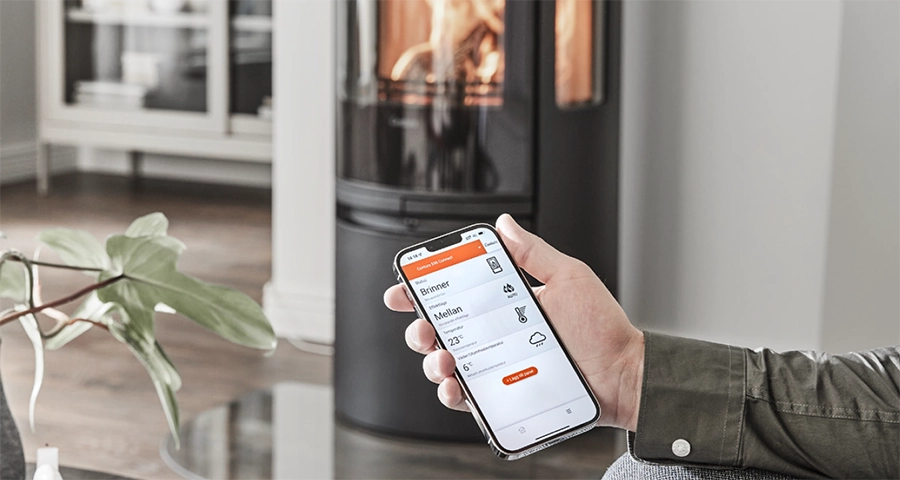 En person håller en smartphone med en app för att styra en eldstad. I bakgrunden syns en modern, brinnande kamin i ett hemtrevligt rum.