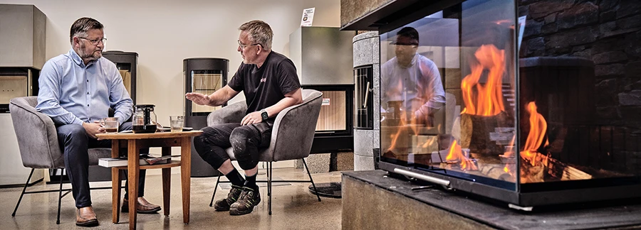 Två män samtalar i ett showroom för eldstäder; en modern kamin med brinnande eld syns i förgrunden.