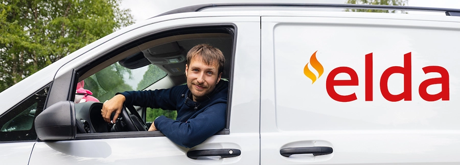 En man sitter i förarsätet på en vit skåpbil med texten "elda" i rött på sidan, omgiven av grönska.