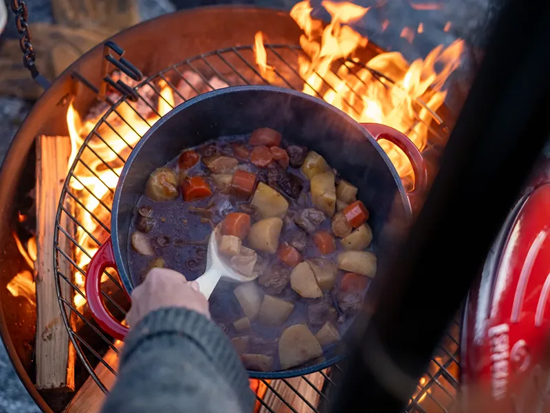 Gryta med kött och grönsaker tillagas över öppen eld på grill. En hand rör om med en slev.