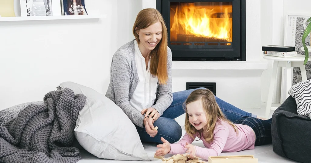 En kvinna och ett barn leker på golvet framför en tänd braskamin i ett modernt vardagsrum.