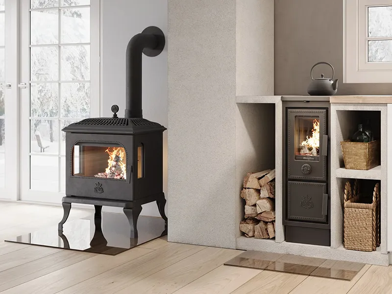 Svart vedeldad kamin i lantligt vardagsrum med stora fönster. Kamin vedförvaring och korgar syns bredvid. Elden brinner klart i kaminen.