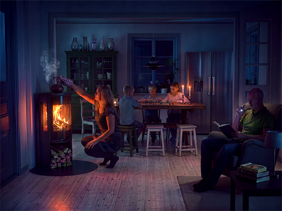 En kvinna lägger ved i en braskamin, medan en familj sitter vid ett bord med tända ljus. Mysig atmosfär i ett vardagsrum.