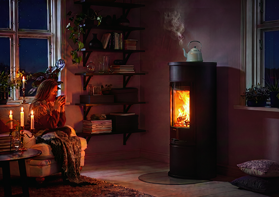 En kvinna sitter på en soffa och dricker något varmt bredvid en brinnande kamin i ett mysigt rum med tända ljus och hyllor.