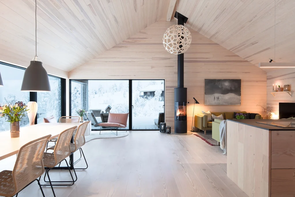 Modernt vardagsrum med ljusa träpaneler, stor kamin och panoramafönster mot snölandskap. Elegant inredning med matplats och soffhörna. Vinterutsikt.