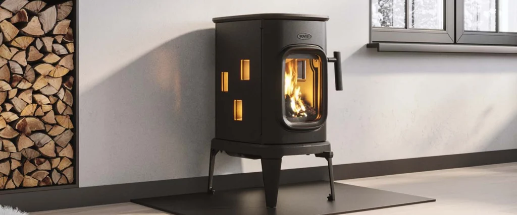 Svart fristående vedkamin i modern design med glaslucka, brinnande eld inuti, placerad på mörk platta. Vedförvaring till vänster mot ljus vägg.