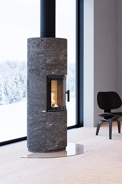En modern, grå stenbeklädd kamin med glasdörr står i ett ljust rum med stora fönster och snöig utsikt. En svart stol syns i bakgrunden.