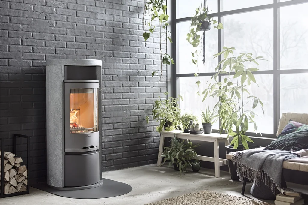 Modern kamin i ett stilrent vardagsrum med grå tegelvägg, omgiven av gröna växter och stora fönster. Eldstadens glasdörr visar en varm eld.