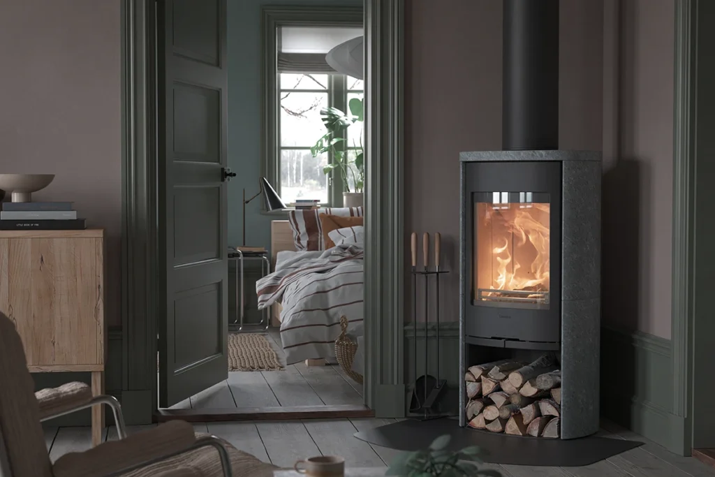 Modernt vardagsrum med en brinnande kamin till höger och en öppning mot ett sovrum med säng och grön växt, inredningsdetaljer synliga.