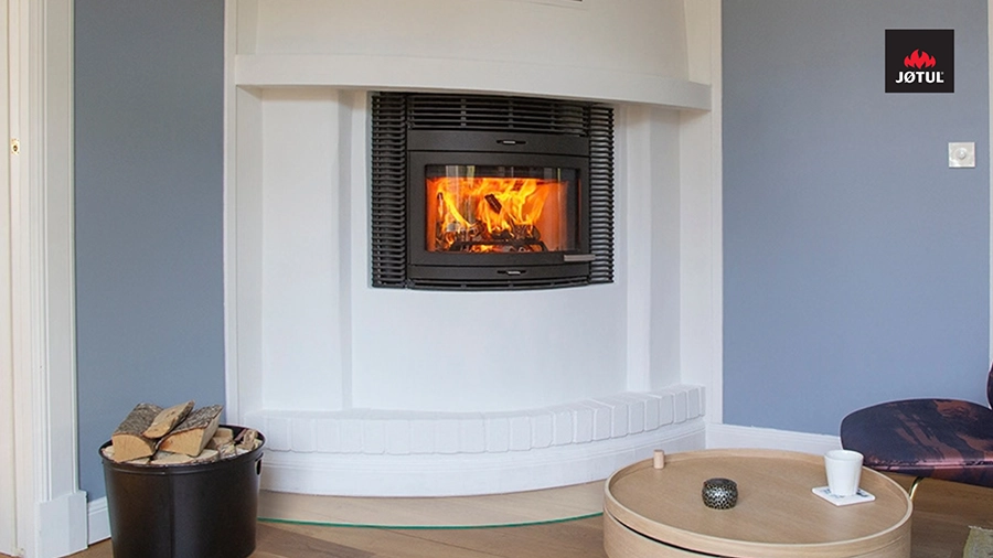 Inbyggd kamin i vit vägg, eld brinner inuti. Vedkorg på golvet, rund träbord med kopp framför. Jøtul-logotyp i övre högra hörnet.