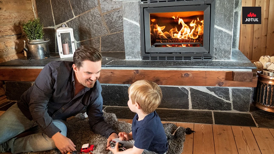 En man och ett barn leker på en matta framför en tänd Jøtul-kamin i ett mysigt vardagsrum med träpaneler och stengolv.