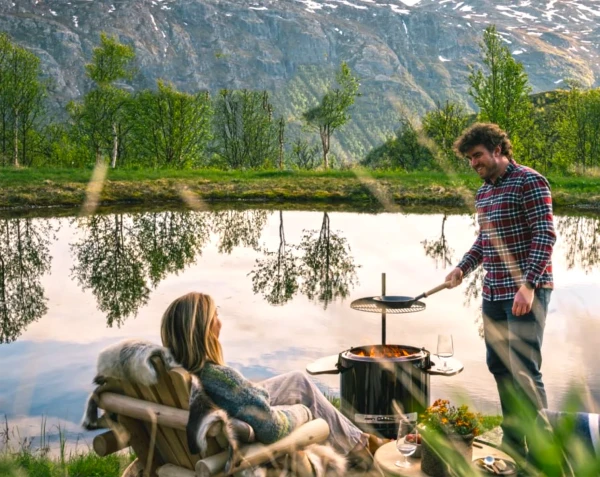 Man grillar vid en sjö, omgiven av natur och berg. Kvinnan sitter i en stol täckt med filt. Avslappnad utomhusmiljö.